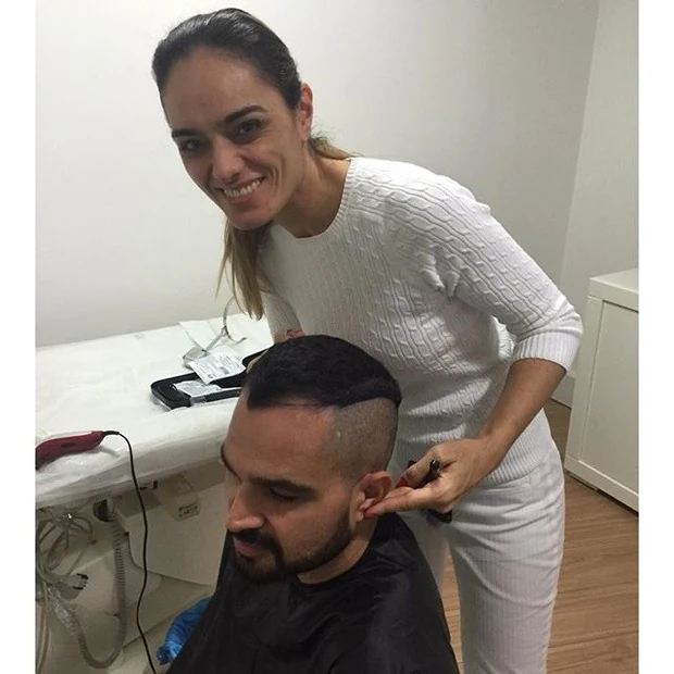 Luciano Camargo sentado na clínica, com laterais do cabelo raspadas e a médica ao lado.