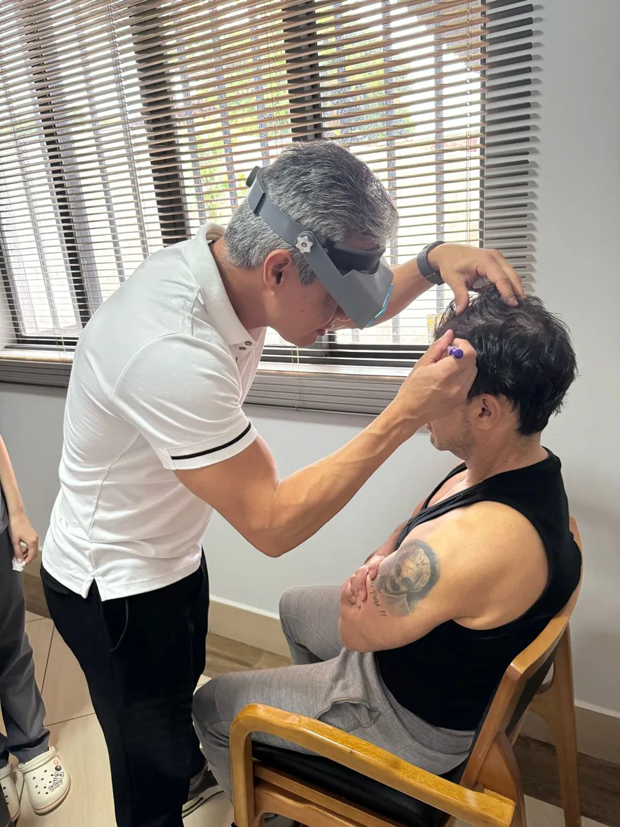 Zezé Di Camargo sentado enquanto o médico marca a linha frontal do cabelo com uma caneta.